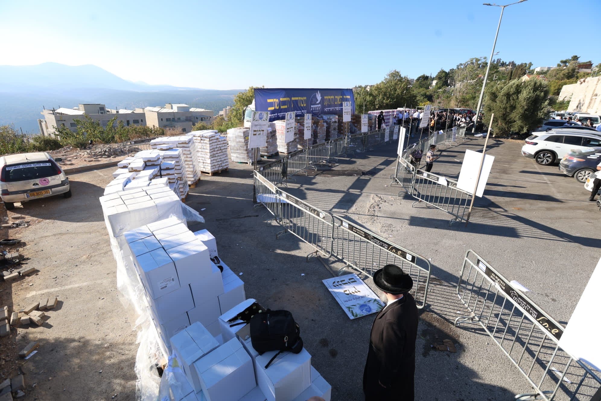 The Yom Tov distribution site on the Tzfat hillside, pallets and crowds under the morning sun
