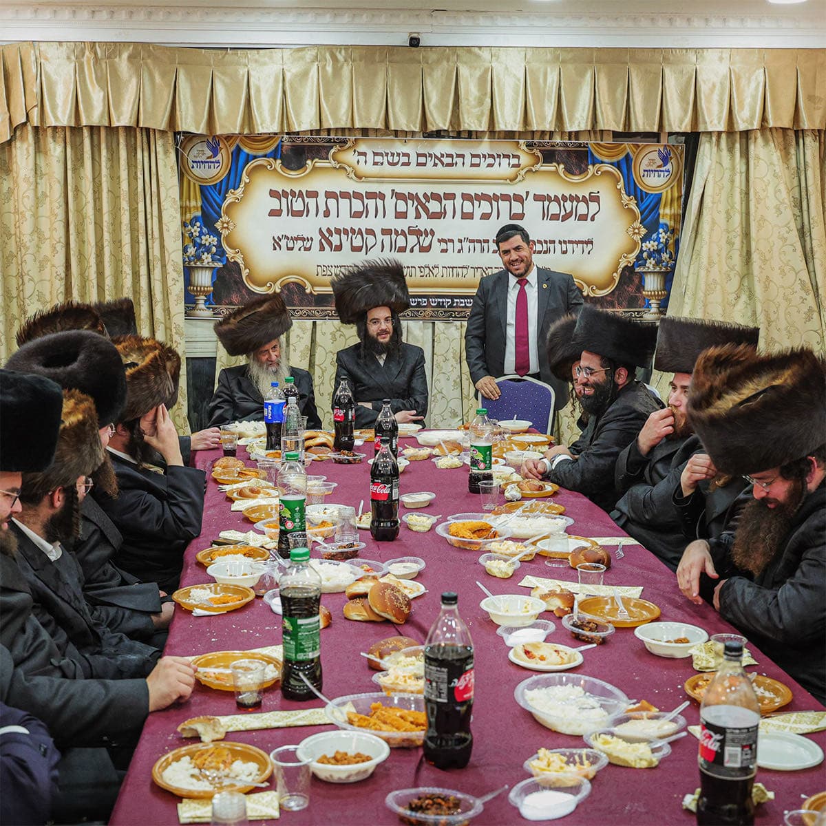 Rabbi Shlomo Katina standing at the head of a long, laden table in Tzfat, addressing rabbanim and guests seated along both sides beneath a Hebrew welcome banner